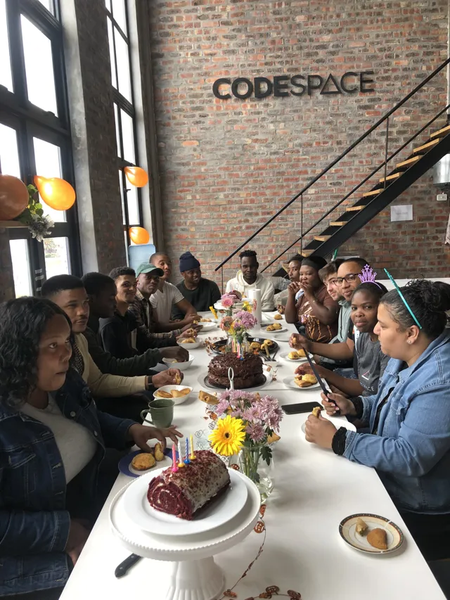 Students sharing birthday cake around the studio table beneath the CodeSpace sign