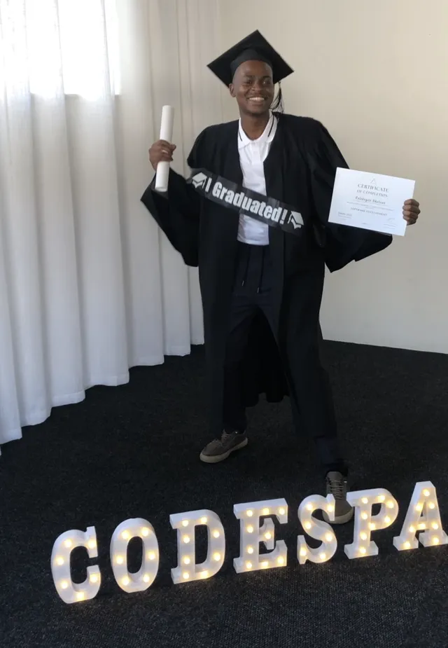 A graduate in cap and gown holding his certificate beside CODESPACE letter lights